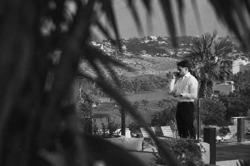 Eliezer al teléfono en una terraza, fotografiado de lejos entre las hojas de una palmera